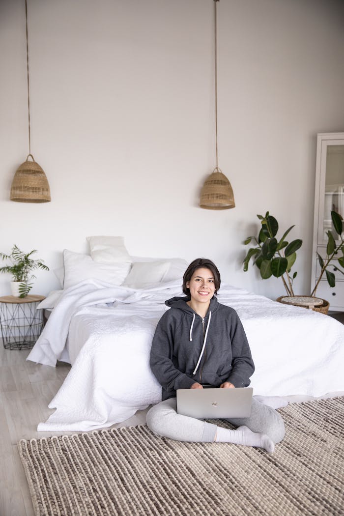 A woman sitting on the floor in a modern, cozy bedroom working on a laptop.