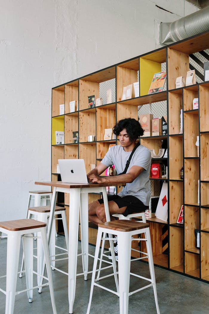 A young man focused on his laptop while sitting in a stylish, modern café with wooden shelving.
