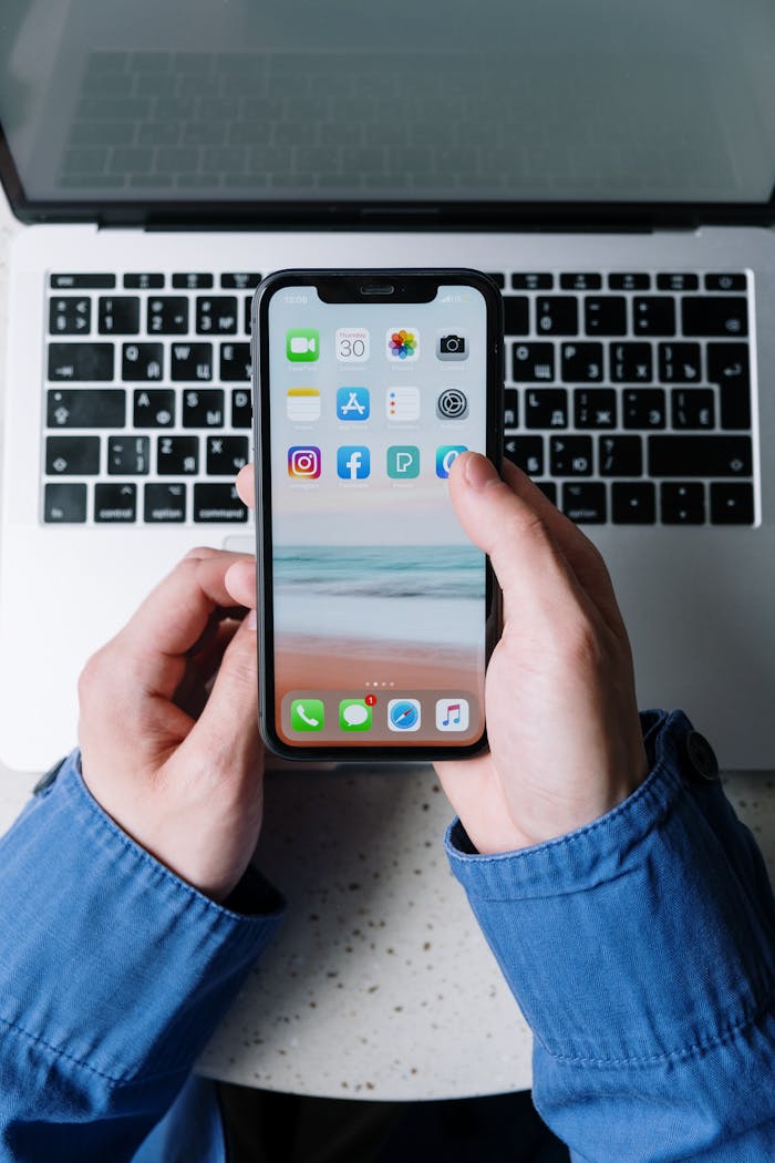 Close-up view of hands holding a smartphone with apps over a laptop keyboard.