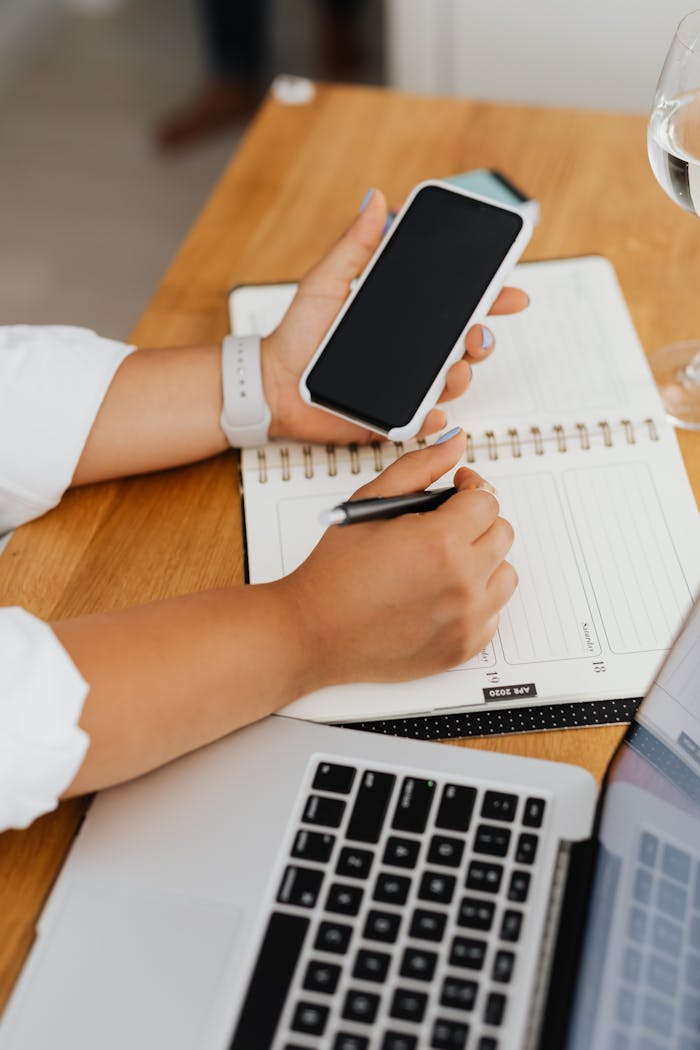 Woman multitasking with a smartphone and writing notes at a workspace.
