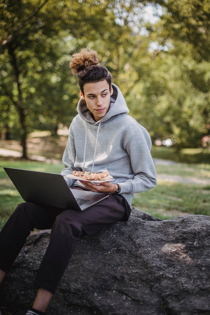 Concentrated male learner sitting on stone and browsing netbook for studies while having lunch with fast food in park