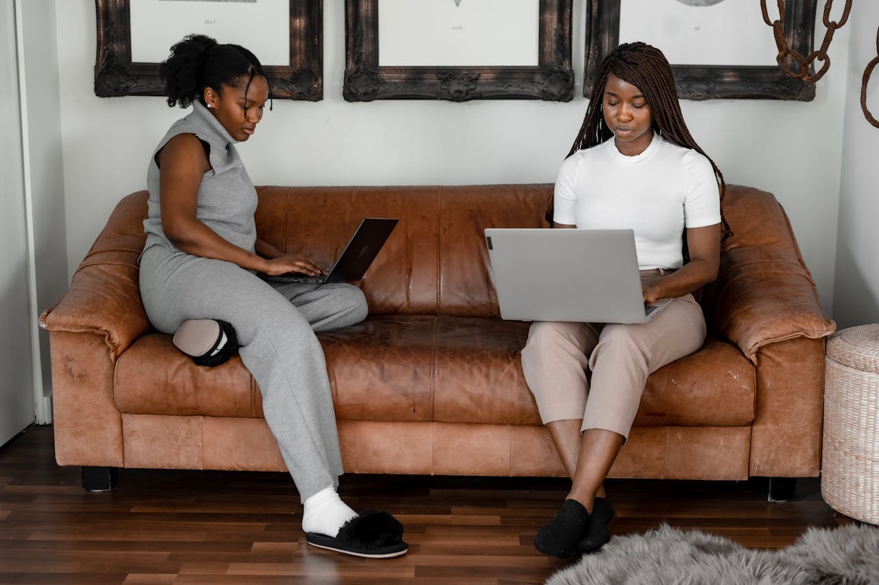 Two African American women sitting on a couch, working from home on laptops.